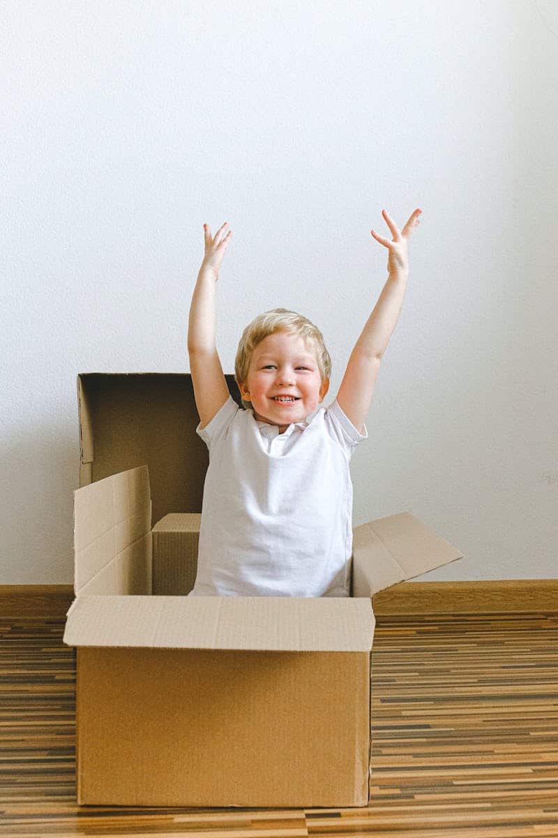Cheerful child playing in a cardboard box at home, arms uplifted in joy, expressing happiness and creativity.