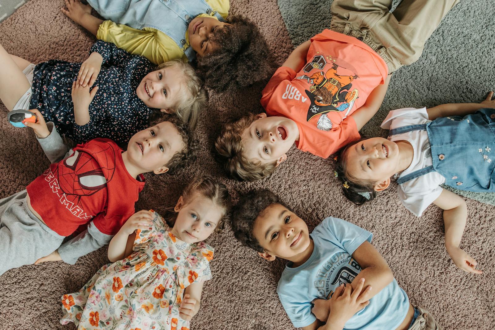 A joyful group of children lying on carpet, forming a circle and smiling at the camera.